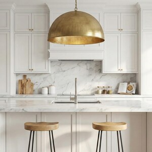 May include: A modern kitchen with white cabinets, a marble countertop, and a large brass pendant light. Two wooden stools with black metal legs sit at the island. The backsplash is marble, and there are wooden cutting boards and jars on the counter.
