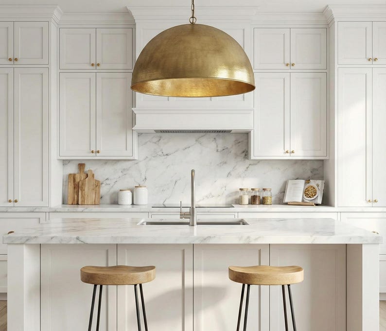 May include: A modern kitchen with white cabinets, a marble countertop, and a large brass pendant light. Two wooden stools with black metal legs sit at the island. The backsplash is marble, and there are wooden cutting boards and jars on the counter.