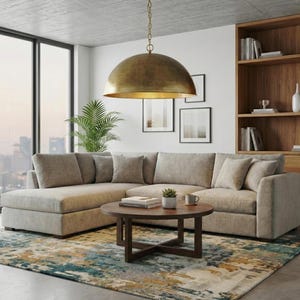 May include: A modern living room featuring a large, light-colored sectional sofa with throw pillows. A round, dark wood coffee table holds books and a small plant. A large, gold-toned pendant light hangs above. A patterned rug and wooden bookshelf complete the scene.
