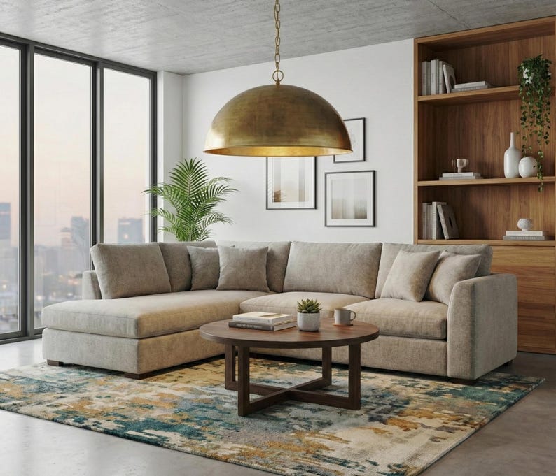 May include: A modern living room featuring a large, light-colored sectional sofa with throw pillows. A round, dark wood coffee table holds books and a small plant. A large, gold-toned pendant light hangs above. A patterned rug and wooden bookshelf complete the scene.