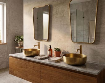 May include: A bathroom vanity featuring a long wooden cabinet with a marble countertop, two round brushed gold vessel sinks, two matching gold faucets, and two asymmetrical wall-mounted mirrors with thin gold frames, plus two small potted plants.
