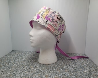May include: A white and pink surgical scrub hat with a purple tie. The hat has a print of a city skyline with pink and purple accents.