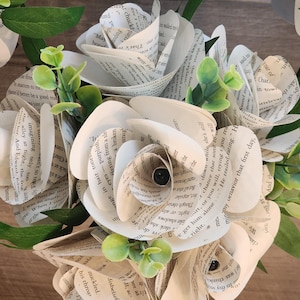 May include: A bouquet of handcrafted paper flowers made from vintage book pages with visible printed text. The arrangement features multiple layered rose-shaped blooms interspersed with small, bright green artificial leaves and stems.