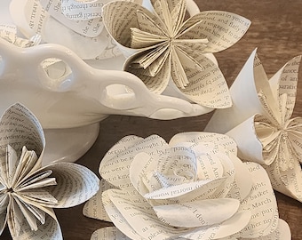 May include: A collection of handcrafted paper flowers made from vintage book pages with visible printed text. The flowers feature various folded petal designs, including layered rose-like shapes and star-like blossoms, arranged around a white ceramic dish.