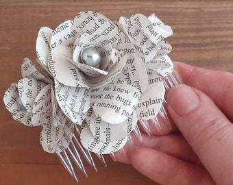 May include: A white paper flower hair comb with a silver pearl in the center. The flower is made from pages of a book and the comb is clear plastic.