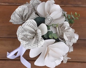 May include: A bouquet of white paper flowers made from book pages. The flowers are arranged in a circular shape and are tied together with a white ribbon. The bouquet is on a wooden surface.