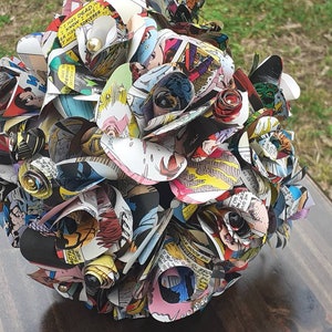May include: A bouquet of paper flowers made from comic book pages. The flowers are arranged in a circular shape and have a variety of colors, including red, blue, yellow, and green. The flowers are decorated with small, shiny beads.