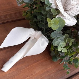 May include: A white book page bouquet with a white satin ribbon. The bouquet is made of paper roses with text from a book. The bouquet is surrounded by green eucalyptus leaves.