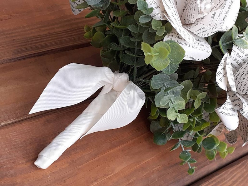May include: A white book page bouquet with a white satin ribbon. The bouquet is made of paper roses with text from a book. The bouquet is surrounded by green eucalyptus leaves.