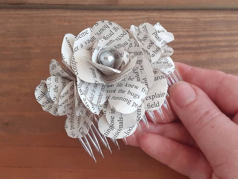 Puede incluir: Un peine para el cabello con forma de flor blanca de papel con una perla plateada en el centro. La flor est&aacute; hecha de p&aacute;ginas de un libro y el peine es de pl&aacute;stico transparente.