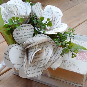 May include: A decorative bouquet of three handcrafted flowers made from printed book pages, featuring white paper petals with black text, accented with small green plastic leaves and stems, resting against a rectangular book with a cream cover.