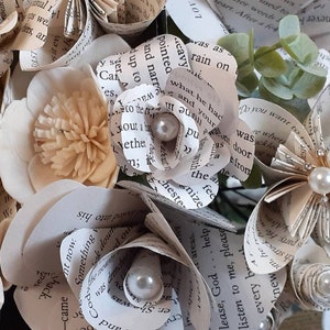 May include: A bouquet of paper flowers made from book pages, with pearl accents. The flowers are in shades of cream and white, and the bouquet is tied with a ribbon.