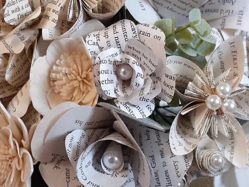 May include: A bouquet of paper flowers made from book pages, with pearl accents. The flowers are in shades of cream and white, and the bouquet is tied with a ribbon.