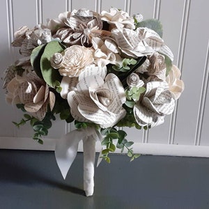 May include: A bouquet of white paper roses made from book pages, with greenery and pearl accents. The bouquet is tied with a white ribbon.