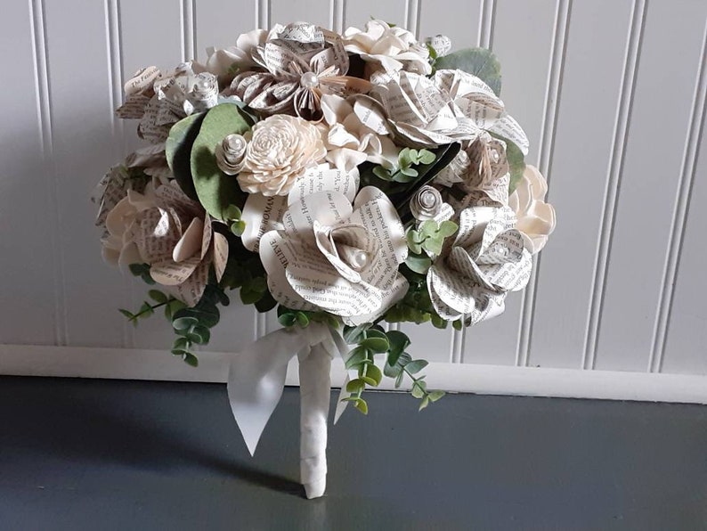 May include: A bouquet of white paper roses made from book pages, with greenery and pearl accents. The bouquet is tied with a white ribbon.