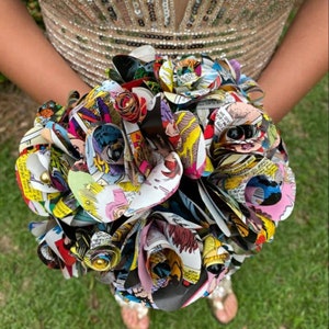 May include: A bouquet of colorful paper roses made from comic book pages. The roses are arranged in a circular shape and are held together with a ribbon. The bouquet is a unique and creative way to celebrate a special occasion.