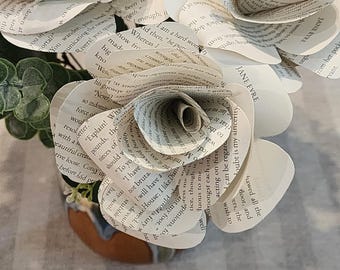 May include: A decorative flower sculpture crafted from pages of the book Jane Eyre, featuring layered paper petals with visible printed text. The flower is accompanied by small green artificial leaves and is set in a rounded base.