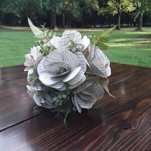 May include: A bouquet of handcrafted flowers made from recycled book pages with printed black text. The arrangement features multiple paper roses interspersed with small green artificial leaves and fern-like foliage, creating a rounded floral structure.