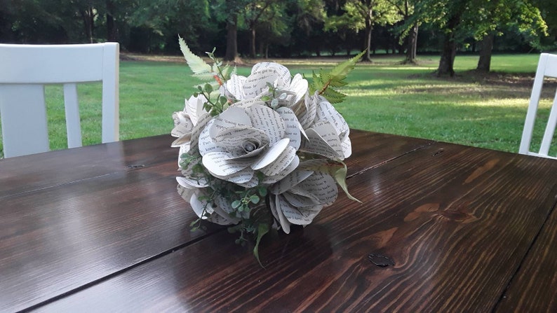 May include: A bouquet of handcrafted flowers made from recycled book pages with printed black text. The arrangement features multiple paper roses interspersed with small green artificial leaves and fern-like foliage, creating a rounded floral structure.