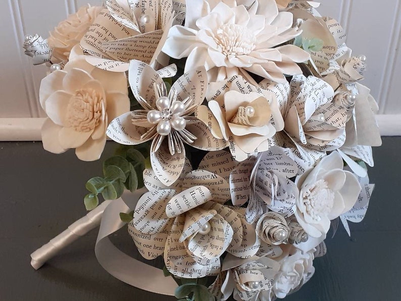 May include: A bouquet of cream-colored paper flowers with a satin ribbon handle. The flowers are made from pages of old books and are accented with pearl beads.