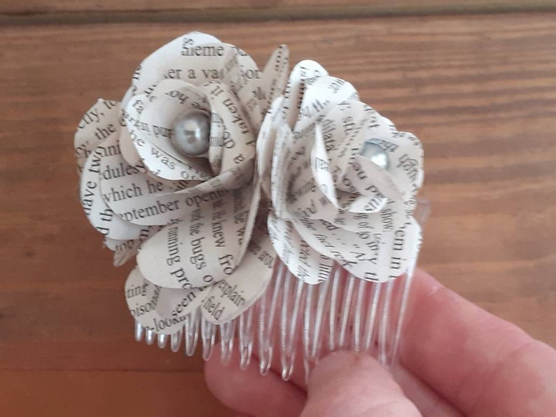 Puede incluir: Un peine para el cabello de flores blancas de papel con dos acentos de perlas. Las flores est&aacute;n hechas de p&aacute;ginas de un libro y est&aacute;n unidas a un peine de pl&aacute;stico transparente.