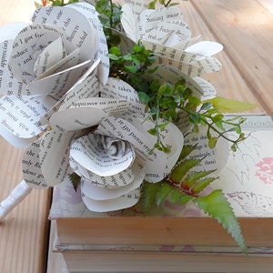 May include: A decorative bouquet of roses crafted from printed book pages with visible black text, accented by small green artificial leaves. The paper flowers are arranged in a cluster and rest upon a stack of two hardcover books with light-colored covers.