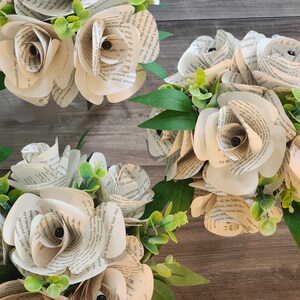 May include: Three bouquets of handcrafted paper roses made from vintage book pages. Each flower features curled petals with visible printed text, accented by small green artificial leaves and sprigs tucked between the paper blooms.