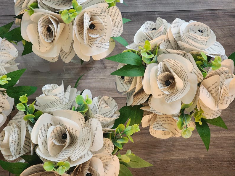 May include: Three bouquets of handcrafted paper roses made from vintage book pages. Each flower features curled petals with visible printed text, accented by small green artificial leaves and sprigs tucked between the paper blooms.