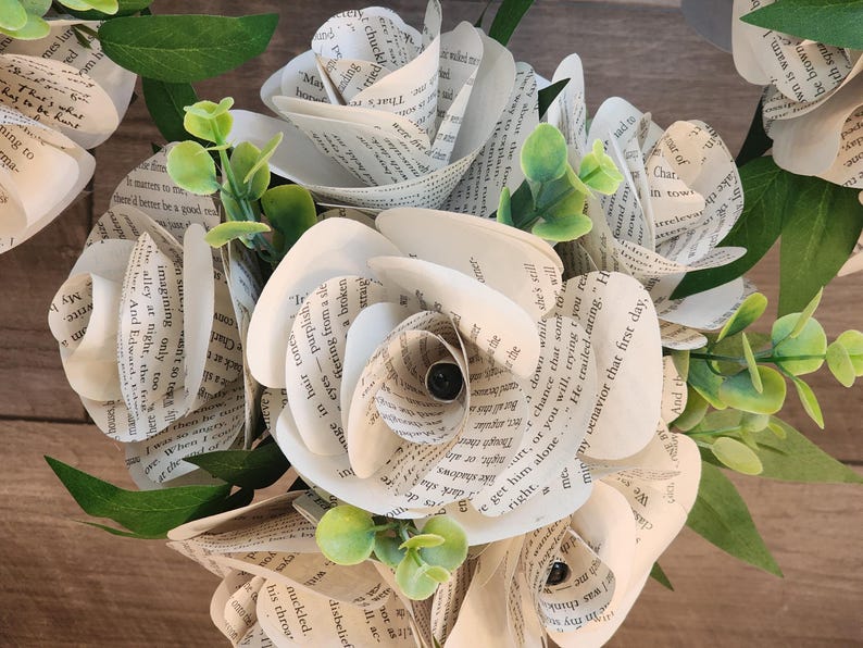 May include: A bouquet of handcrafted paper flowers made from vintage book pages with visible printed text. The arrangement features multiple layered rose-shaped blooms interspersed with small, bright green artificial leaves and stems.