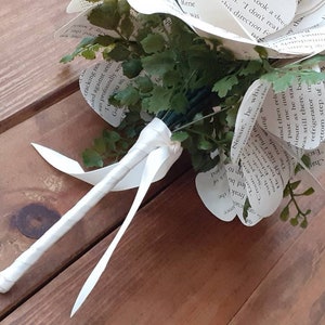 May include: A bouquet of flowers crafted from printed book pages with visible black text. The arrangement includes green artificial foliage interspersed among the paper petals. The stems are bound together with a white ribbon wrapped around them.