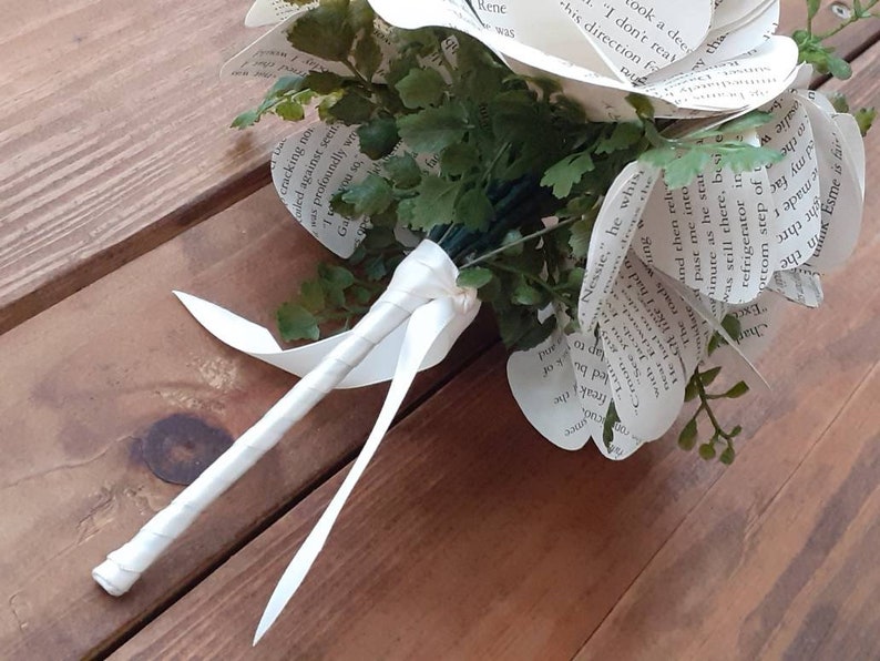 May include: A bouquet of flowers crafted from printed book pages with visible black text. The arrangement includes green artificial foliage interspersed among the paper petals. The stems are bound together with a white ribbon wrapped around them.