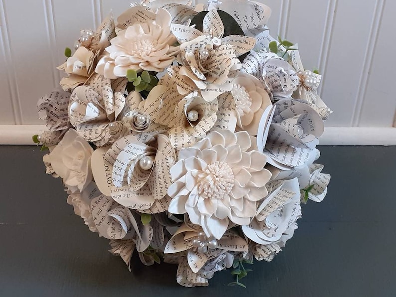 May include: A bouquet of cream-colored paper flowers made from book pages. The bouquet is decorated with small white pearls and green leaves. The text "LOVE NO" is visible on one of the pages.