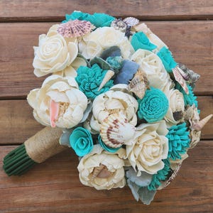May include: A bouquet of cream and turquoise flowers crafted from wood, accented with various seashells including scallop and spiral shells. The stems are bundled together and wrapped in natural tan jute twine, creating a coastal-themed arrangement.