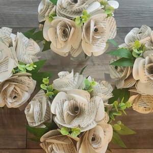 May include: Four bouquets of handcrafted paper flowers made from vintage book pages. Each bouquet features multiple roses with curled petals displaying printed text, accented by small green plastic leaves and stems arranged in clusters.