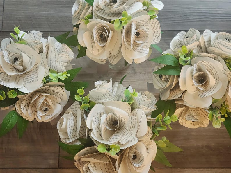 May include: Four bouquets of handcrafted paper flowers made from vintage book pages. Each bouquet features multiple roses with curled petals displaying printed text, accented by small green plastic leaves and stems arranged in clusters.