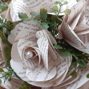 May include: A bouquet of handcrafted paper roses made from printed book pages, featuring curled petals with visible black text. Each rose is accented with a small, round, white faux pearl at its center, interspersed with sprigs of green artificial foliage.