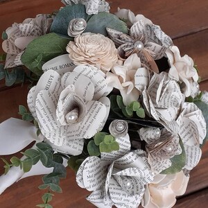 May include: A bouquet of paper flowers made from book pages, with white and cream colored flowers, green leaves, and pearl accents. The bouquet has a white ribbon handle and the text "BELIEVE" and "DARIE ROMAN" are visible on the pages.