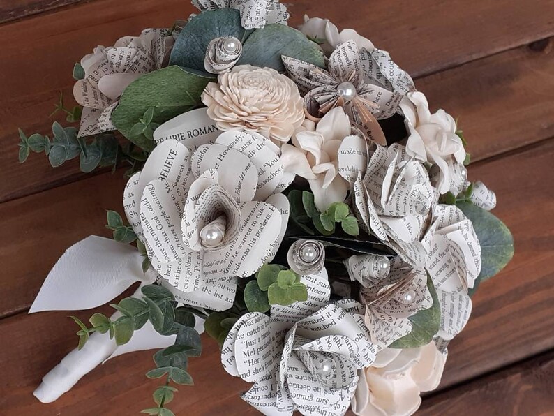 May include: A bouquet of paper flowers made from book pages, with white and cream colored flowers, green leaves, and pearl accents. The bouquet has a white ribbon handle and the text "BELIEVE" and "DARIE ROMAN" are visible on the pages.