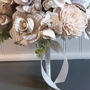 May include: A bouquet of paper flowers made from book pages, with a single white wooden flower and greenery. The bouquet is tied with a white satin ribbon.