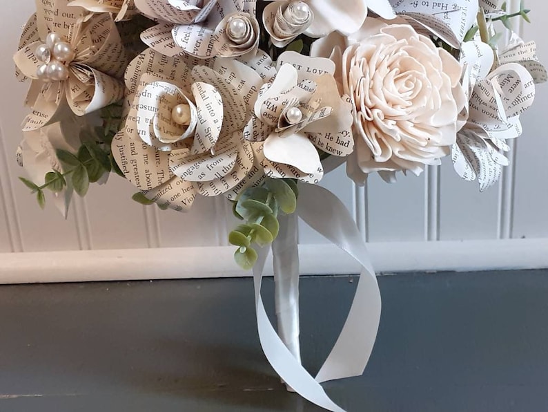 May include: A bouquet of paper flowers made from book pages, with a single white wooden flower and greenery. The bouquet is tied with a white satin ribbon.