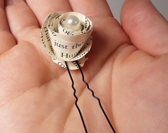 May include: A small white paper rose with a pearl in the center, attached to two black bobby pins. The rose is made from the pages of a book.