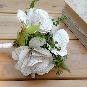 May include: A bouquet of flowers crafted from printed book pages with visible black text. The flowers feature layered paper petals, interspersed with small green plastic leaves and stems. The handle is wrapped in a smooth white satin ribbon.