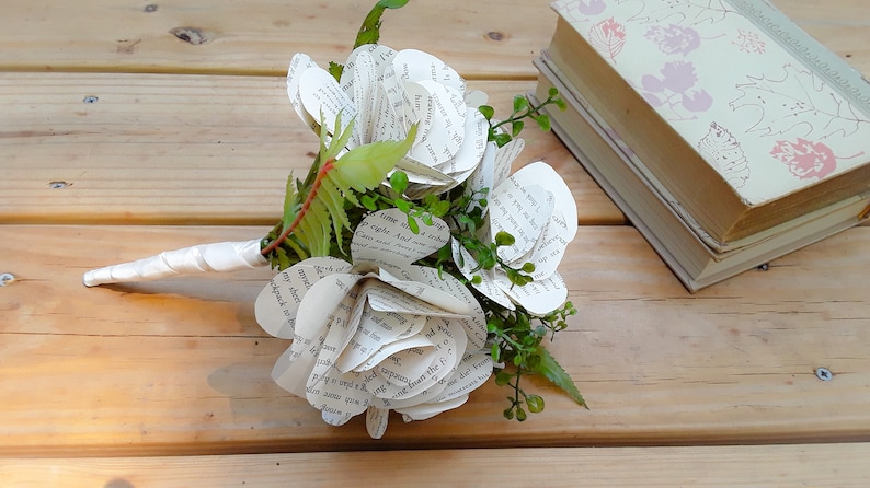 May include: A bouquet of flowers crafted from printed book pages with visible black text. The flowers feature layered paper petals, interspersed with small green plastic leaves and stems. The handle is wrapped in a smooth white satin ribbon.