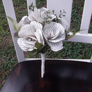 May include: A bouquet of handcrafted flowers made from recycled book pages with printed text. The flowers feature layered paper petals and are accented with small green artificial leaves. The stems are wrapped in a white ribbon to form a handle.