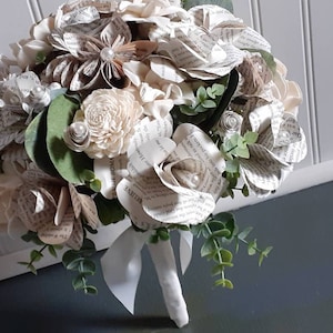 May include: A bouquet of paper flowers made from book pages, with white and cream paper roses, and greenery. The bouquet is tied with a white ribbon and has a white stem.