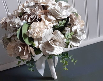 May include: A bouquet of paper flowers made from book pages, with white and cream paper roses, and greenery. The bouquet is tied with a white ribbon and has a white stem.