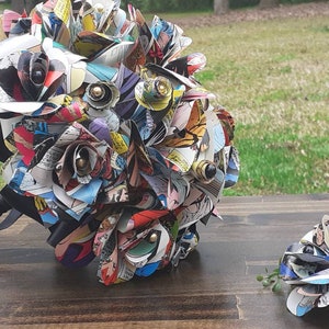 May include: A bouquet of comic book paper roses with black ribbon and a smaller matching boutonniere. The roses are made from colorful comic book pages with black and gold accents.
