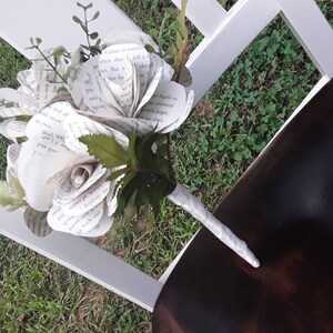 May include: A bouquet of flowers crafted from printed book pages, featuring white paper petals with visible black text. The arrangement includes green artificial leaves and stems, with a handle wrapped in white ribbon and a small pearl accent.
