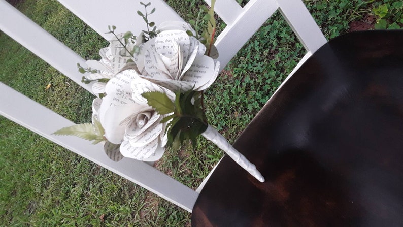 May include: A bouquet of flowers crafted from printed book pages, featuring white paper petals with visible black text. The arrangement includes green artificial leaves and stems, with a handle wrapped in white ribbon and a small pearl accent.