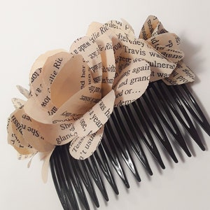 Puede incluir: Un peine de pelo negro con una flor de papel hecha de p&aacute;ginas de libro. La flor est&aacute; hecha de varias capas de papel, cada una con texto impreso. El texto incluye las palabras "Cape Rindu Travis" y "alie Ric".